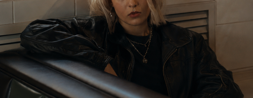 A person with platinum blonde hair sits in a dark leather booth in a diner-like setting, wearing a black leather jacket and black top, with orange and white walls behind them.